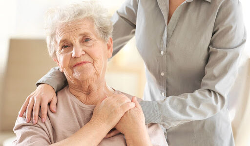 Older woman sitting down holding the hands of another person standing in the background